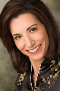 A person with shoulder-length brown hair smiles while wearing a black and gold patterned top.