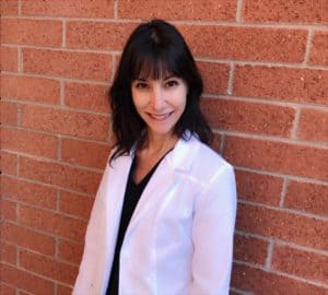 A woman in a white lab coat stands against a brick wall, smiling at the camera.