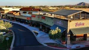 Aerial view of a shopping center with various store signs and a backdrop of mountains under a clear sky.